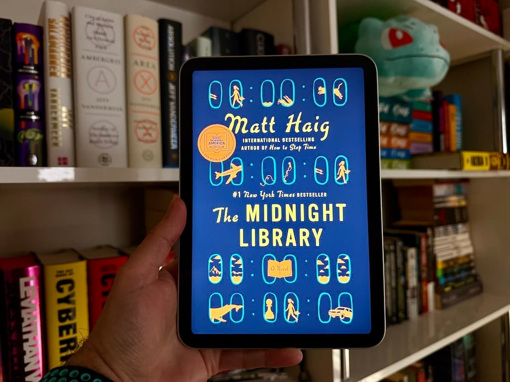 An iPad mini with the cover art for Matt Haig's The Midnight Library being held up against a background of a bookshelf.